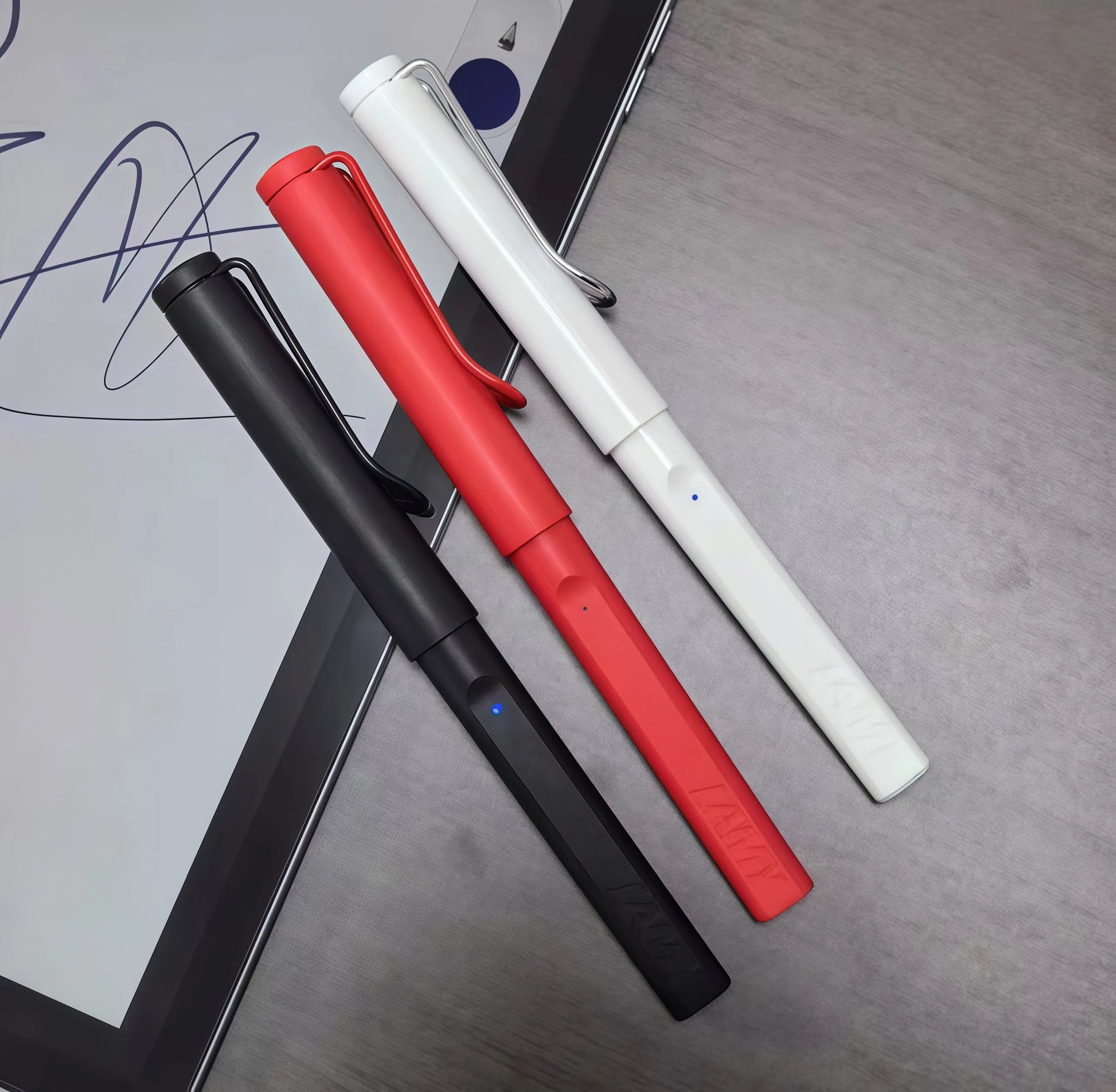 Close-up of LAMY Safari Note+ digital pens in black, red, and white with caps on, arranged diagonally over an iPad screen displaying cursive handwriting. Designed for natural grip, precision, and everyday use.
