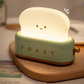 Green toaster night light on wooden chopping board with cutlery, lit toast slice with smiley face emitting warm light